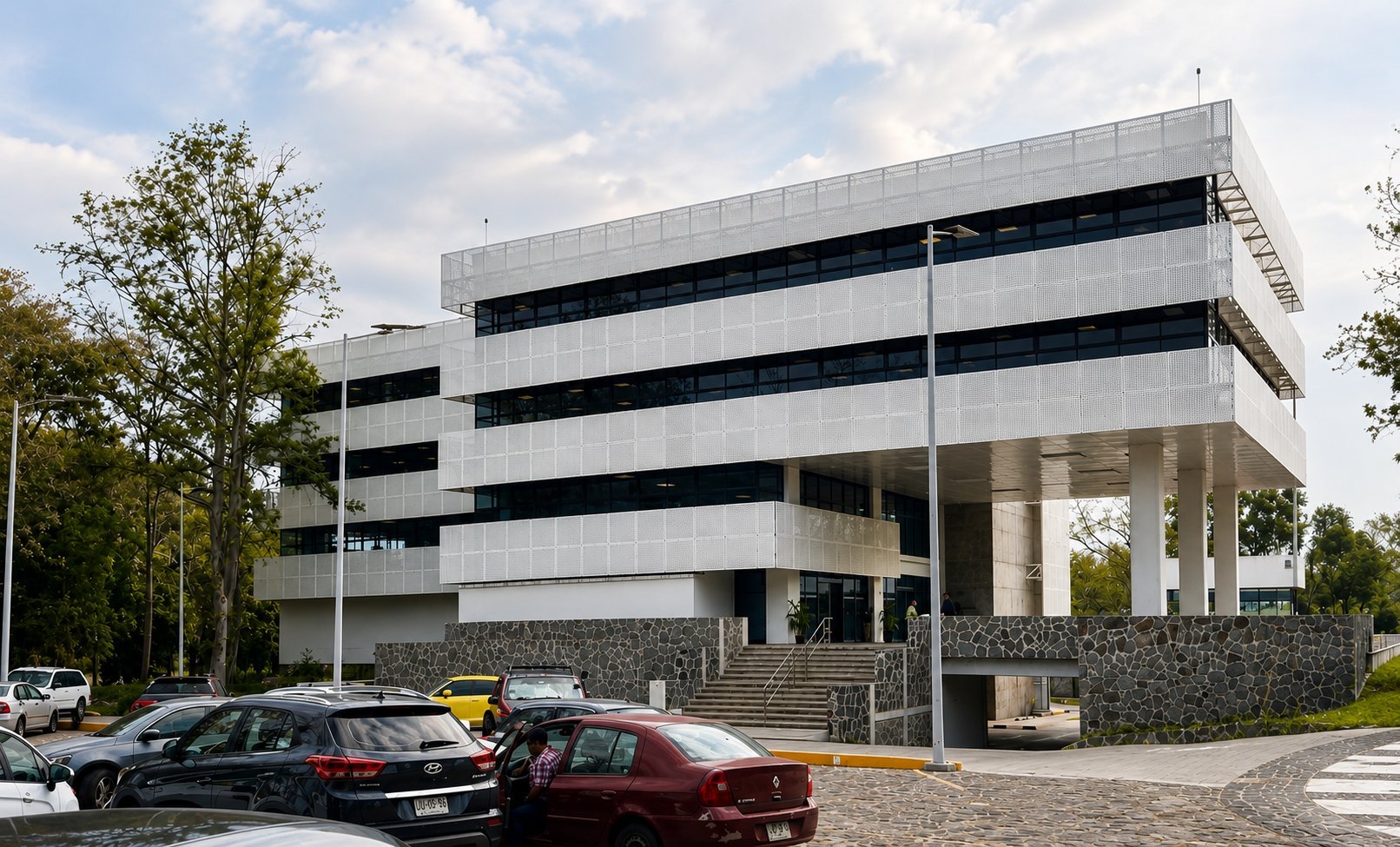 Modern white and black multi-story building with horizontal bands and concrete supports, surrounded by trees and parked cars